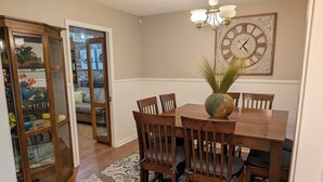 Comedor con mesa de madera, seis sillas, un florero con plantas y un reloj de pared.