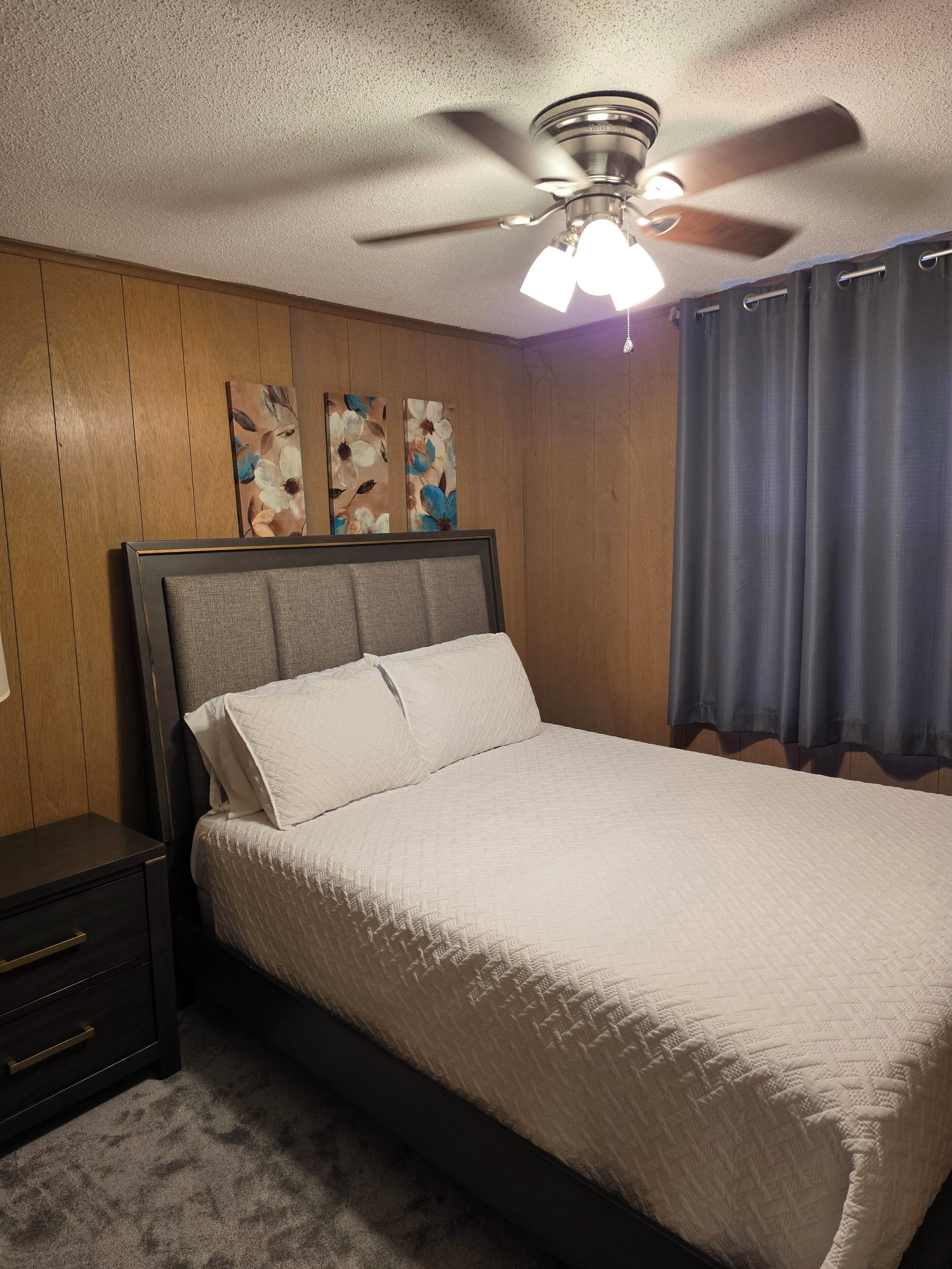 Ein Schlafzimmer mit einem Bett, einem Nachttisch, einem Deckenventilator und Wandbildern.