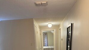 Un pasillo con piso de madera, una puerta que da a un dormitorio y una lámpara fijada en la pared.