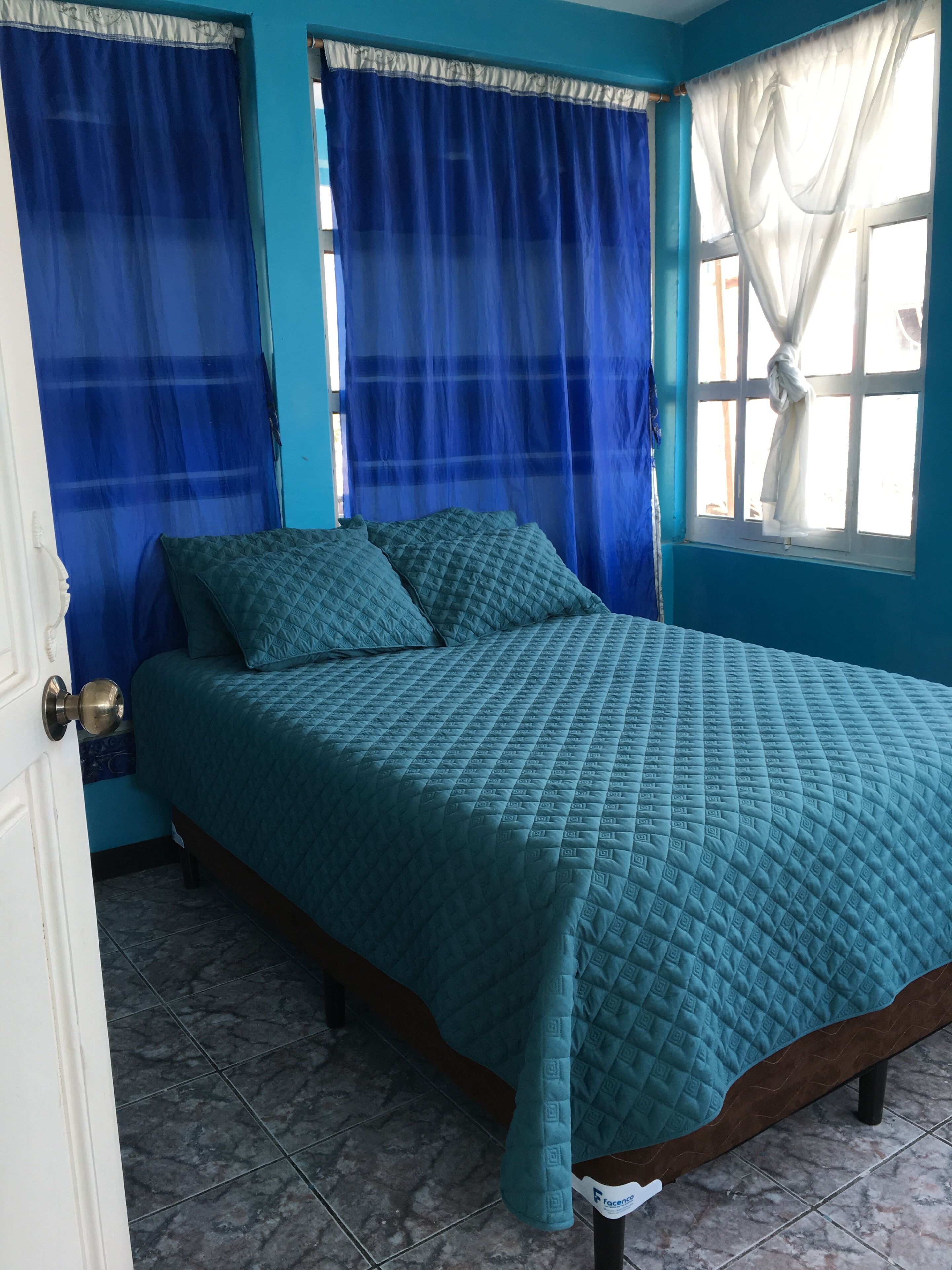 Une chambre à coucher avec une couvre-lit turquoise, des rideaux bleus et une fenêtre ornée d’un rideau blanc.