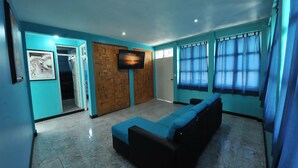 Una habitación con un sofá negro largo, un televisor montado en una pared de madera y una puerta que da a otra estancia.