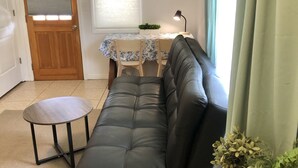 Una sala de estar con un sofá de cuero negro, una pequeña mesa redonda, una mesa de comedor con sillas y un ventanal con cortinas.