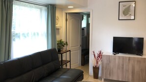 Una sala de estar con un sofá de cuero negro, una mesa de centro redonda, un televisor sobre un soporte de madera y un ventanal con cortinas.