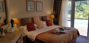 A bedroom with a bed, pillows, a nightstand, a lamp, and a view of a garden through a window. - Hillscapes (Port Alfred)
