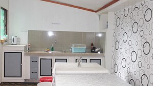 A compact kitchen with a sink, countertop, and cabinets. There is a bed with a white cover in the foreground. - J's Backpackers Hostel (Davao)