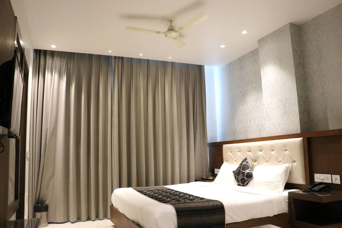 Habitació d'hotel amb llit, tauleta de nit, telèfon, ventilador de sostre i finestra amb cortines.