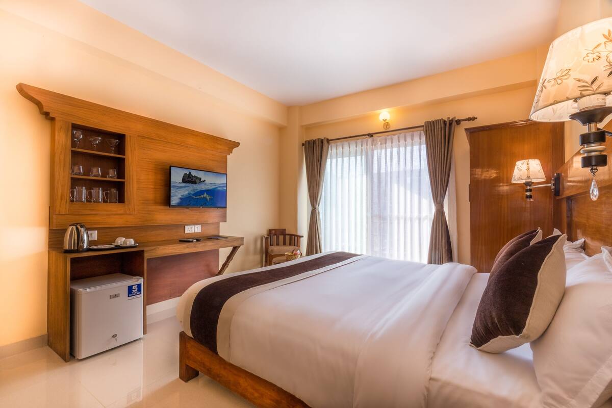 A hotel room with a bed, a wooden cabinet with glassware, a television, a desk, and a small refrigerator.