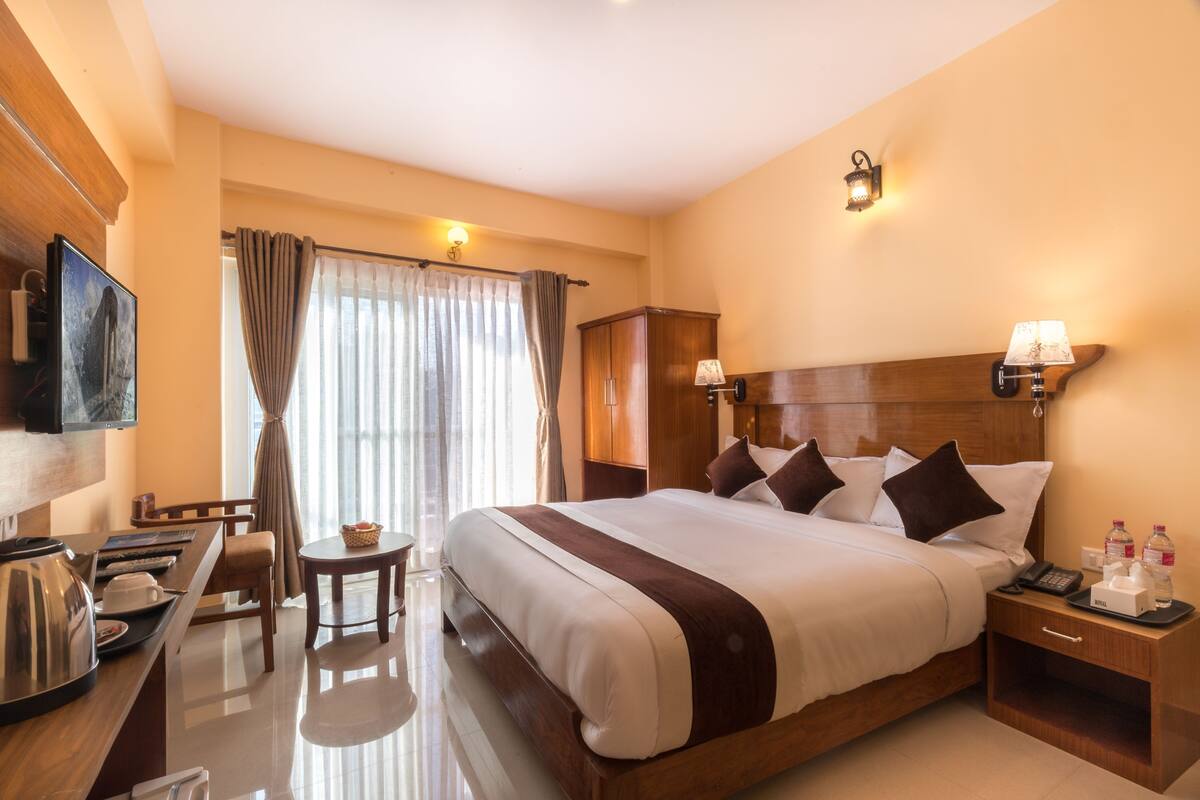 A hotel room with a large bed, bedside tables, a desk with a chair, a television, and a window with curtains.