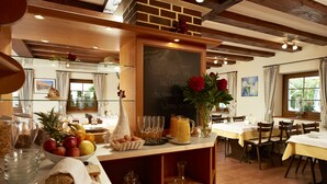 Un bufé de desayuno con una variedad de frutas, pan y bebidas, en un salón con vigas de madera y un menú en una pizarra.