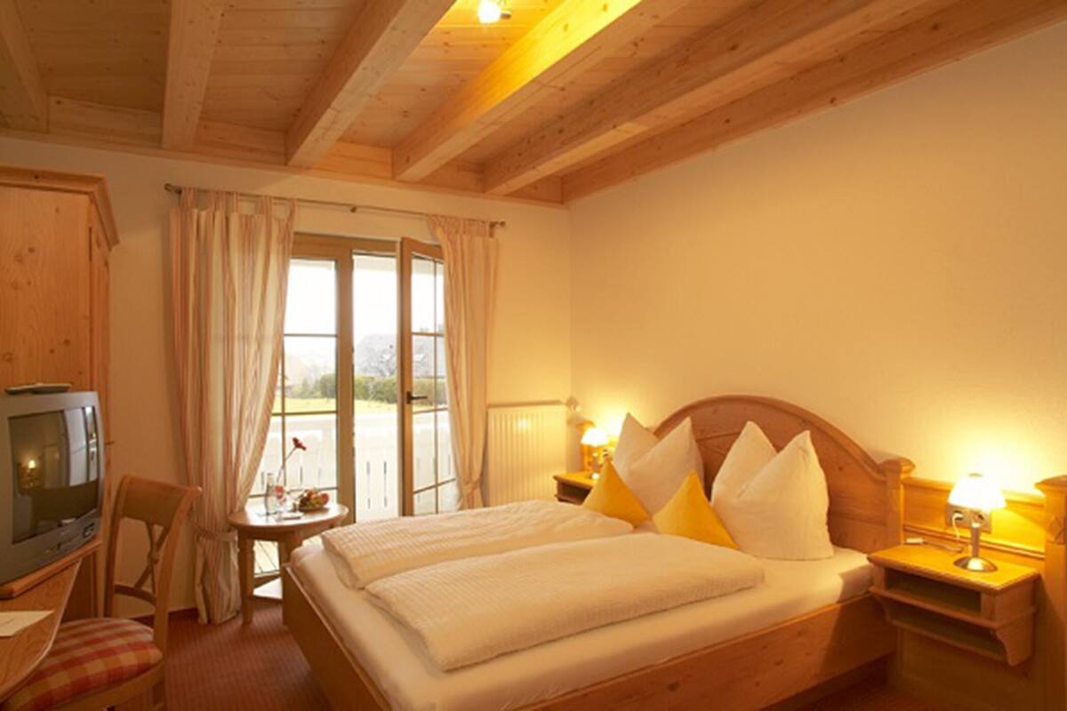 Une chambre d’hôtel confortable avec un plafond en bois, un lit avec une literie blanche et des oreillers jaunes, une table de chevet avec une lampe, une petite table avec une chaise et un téléviseur posé sur un meuble en bois.
