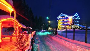 Une route enneigée menant à un bâtiment aux nombreuses fenêtres illuminées, doté de plusieurs balcons et d’un toit recouvert de neige.