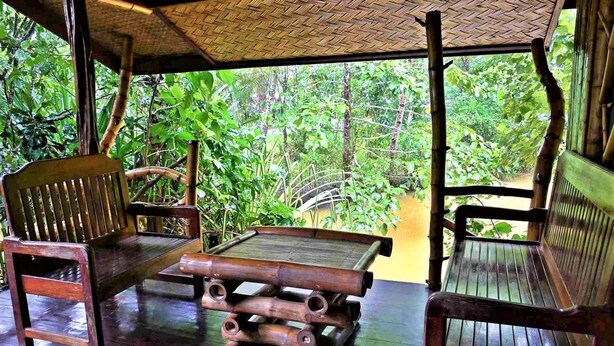 A covered wooden deck with two wooden chairs and a table, surrounded by lush greenery. - Dabdab Tourist Inn (Puerto Princesa)
