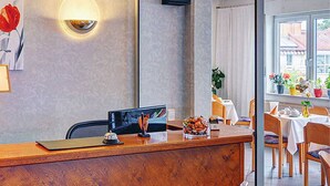 A reception desk with a computer monitor, a small decorative item, and a bowl of snacks. There are chairs and a table with a white tablecloth in the background. - Hotel Post Rosbach (Rosbach vor der Hoehe)