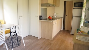 Une cuisine compacte avec des meubles en bois, un four à micro-ondes et un réfrigérateur. On y trouve un coin repas avec une table et des chaises blanches, ainsi qu’un sac à motifs posé sur l’une des chaises.