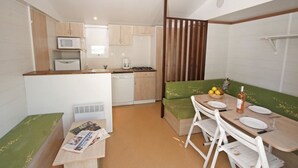 A compact kitchen with a microwave, stove, and sink. There is a dining area with a table and chairs, and a green cushioned bench. - Camping Domaine De Chanteraine (Aiguines)