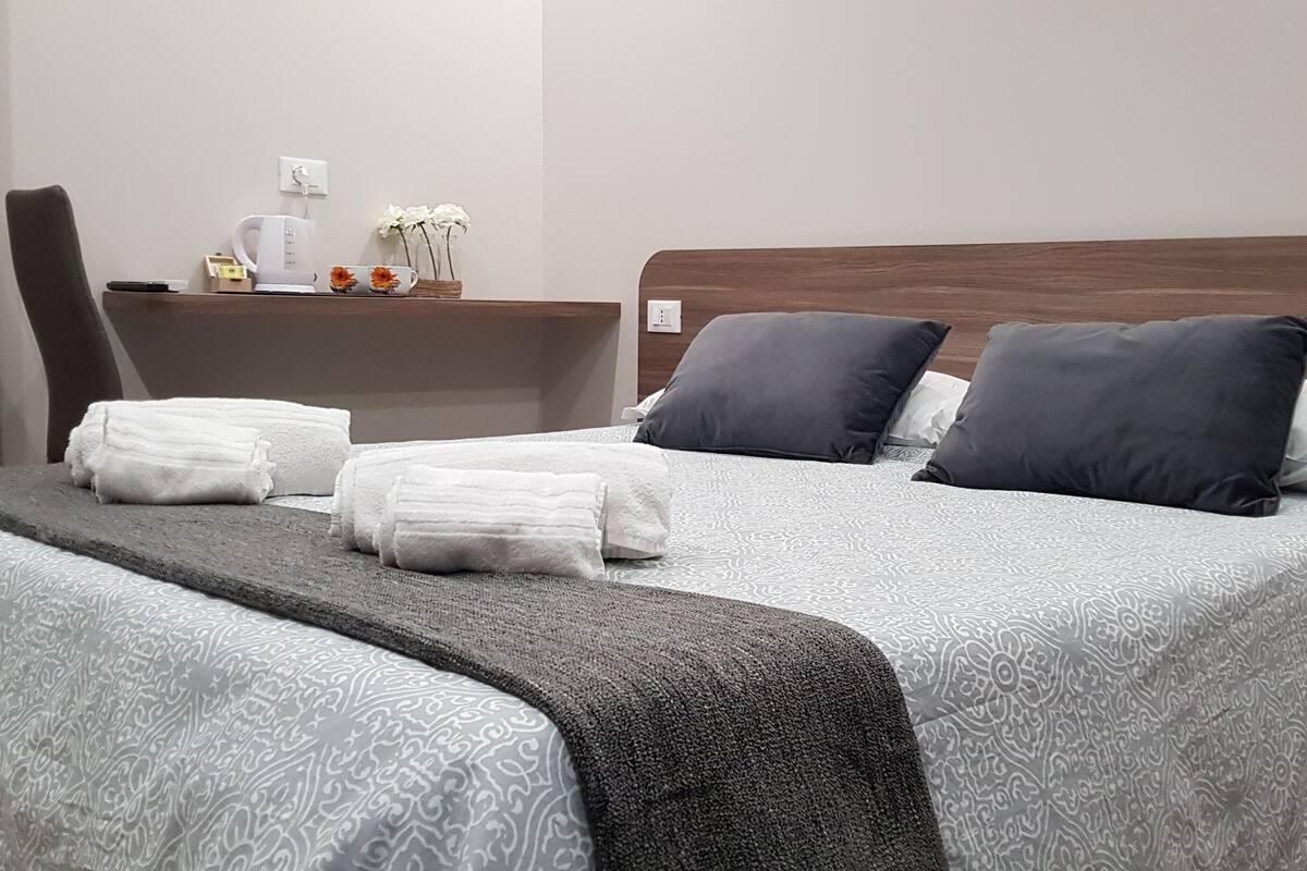 A neatly made bed with a patterned bedspread, two pillows, and a bedside table with a remote control, a phone, and a vase with flowers.