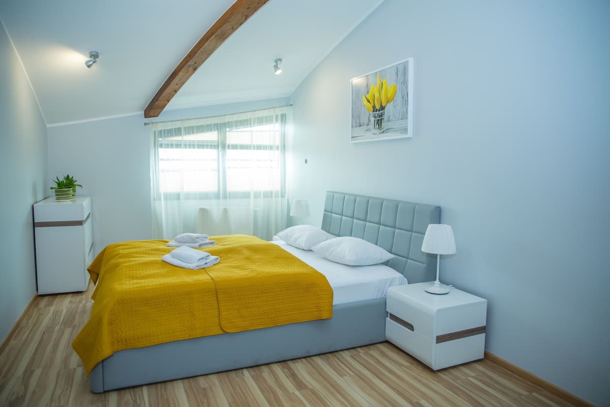 Ein Schlafzimmer mit Bett, Nachttisch, Lampe, Fenster, Vorhängen und einem an der Wand befestigten Bild.