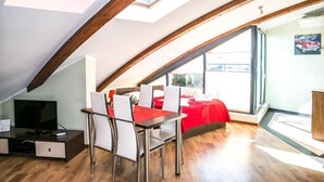 Habitació amb sostre inclinat, zona de menjador amb taula i cadires, televisió i llit amb edredó vermell.