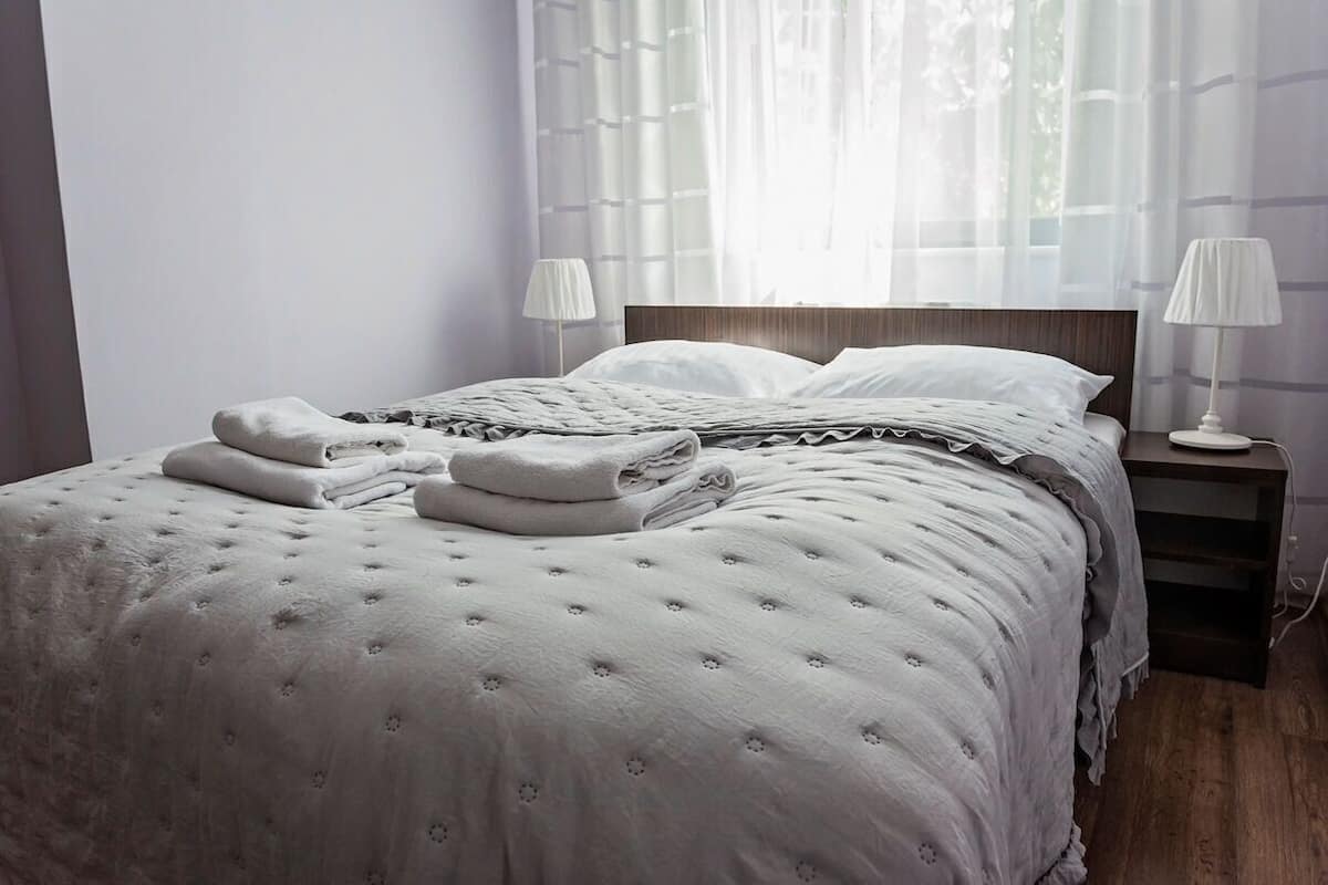 Ein ordentlich bezogenes Bett mit Daunendecke, Kissen und gefalteten Handtüchern. Auf den hölzernen Nachttischen stehen jeweils zwei Nachttischlampen.
