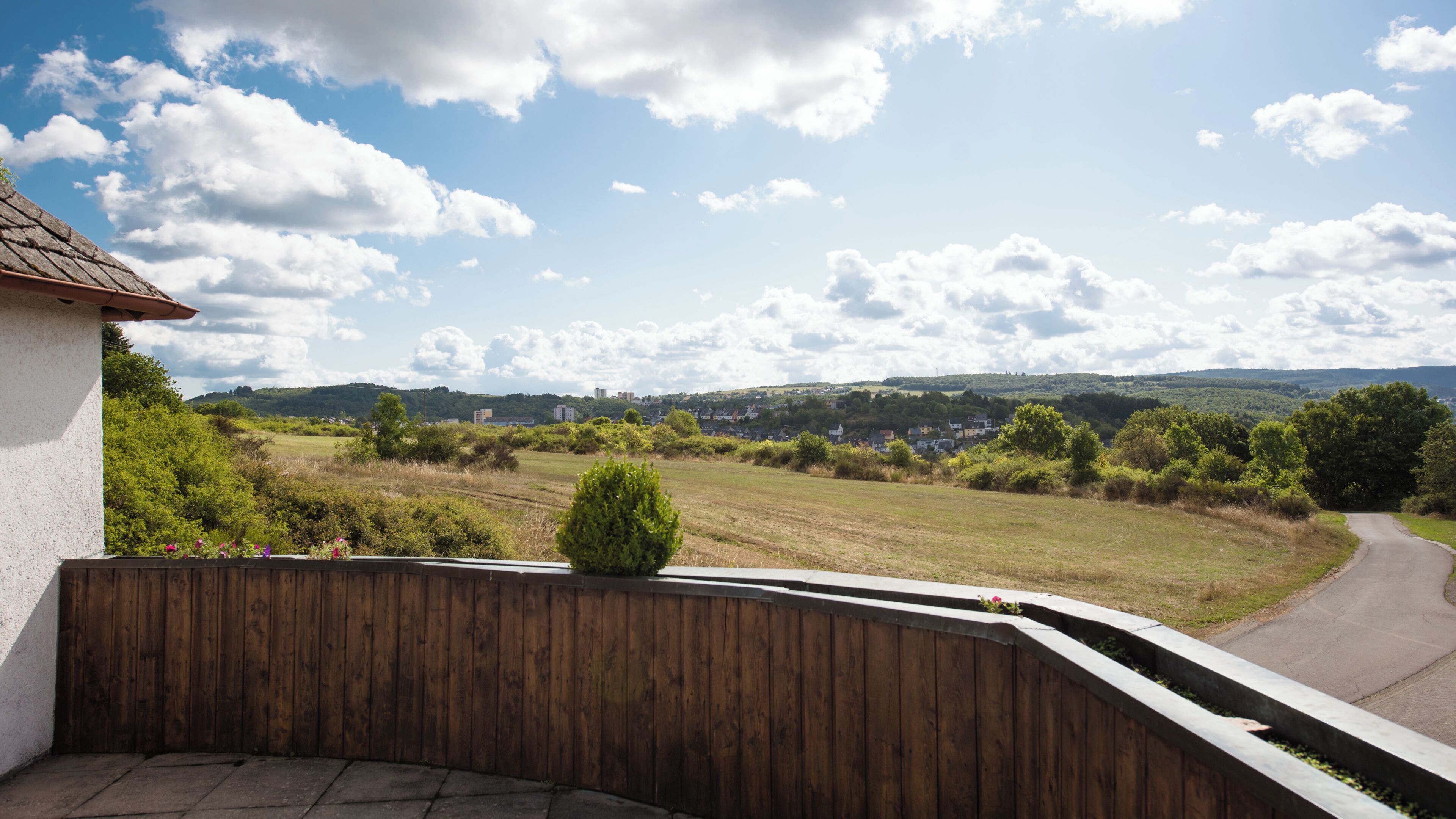 A view from a balcony overlooking a rural landscape with a curved road, greenery, and distant buildings.