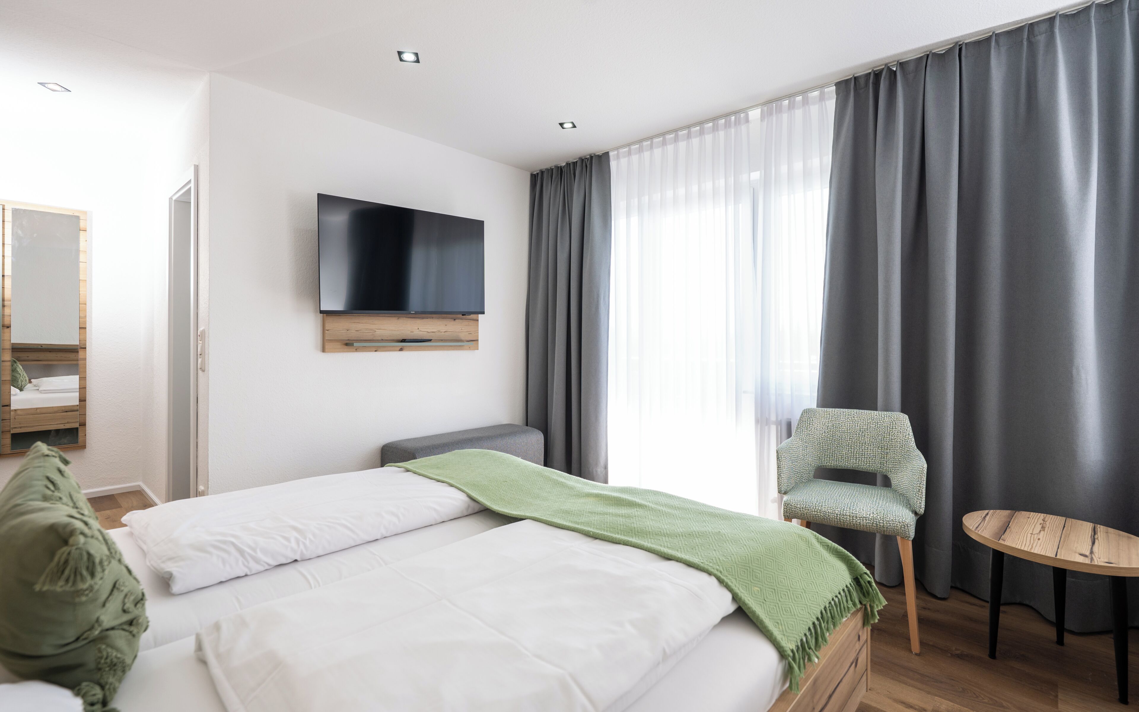 Ein Hotelzimmer mit Bett, Wandfernseher, Stuhl, einem kleinen Tisch und einem Fenster mit Vorhängen.