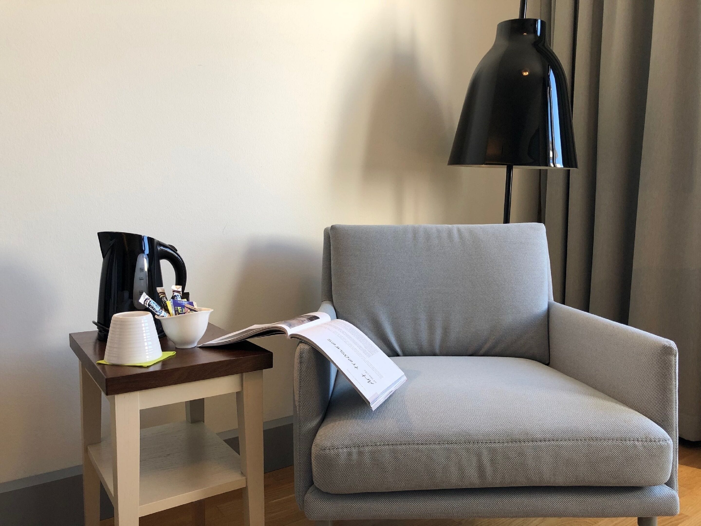 Un sillón pequeño junto a una mesita de madera con una lámpara negra, una taza blanca y un libro.
