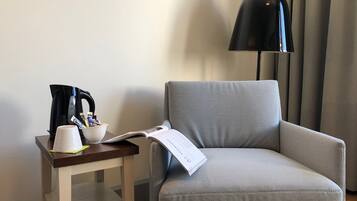 Un sillón pequeño junto a una mesita de madera con una lámpara negra, una taza blanca y un libro.