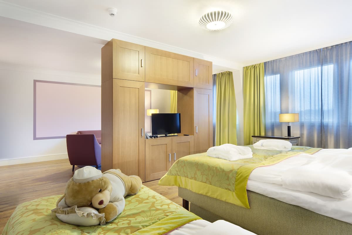 Habitación de hotel con dos camas, televisión, escritorio con lámpara y un oso de peluche sobre una almohada.