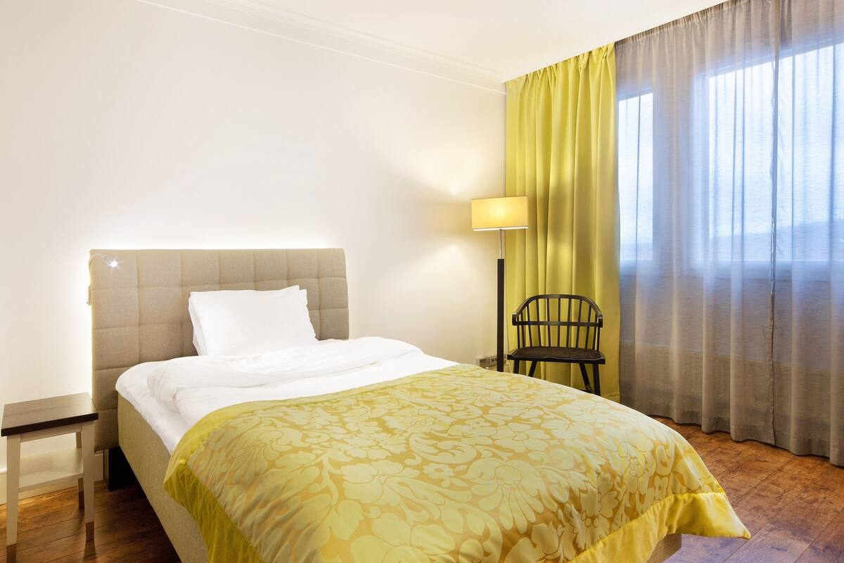 Habitación de hotel con cama, mesita de noche, lámpara, silla y ventana con cortinas.