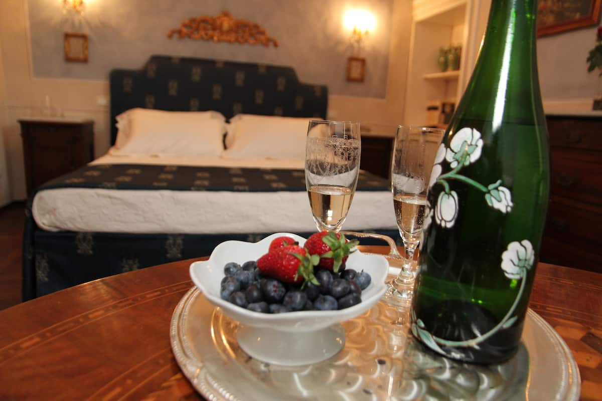 A room with a bed, a dining table with a bowl of fruit and two glasses of wine, and a decorative bottle.