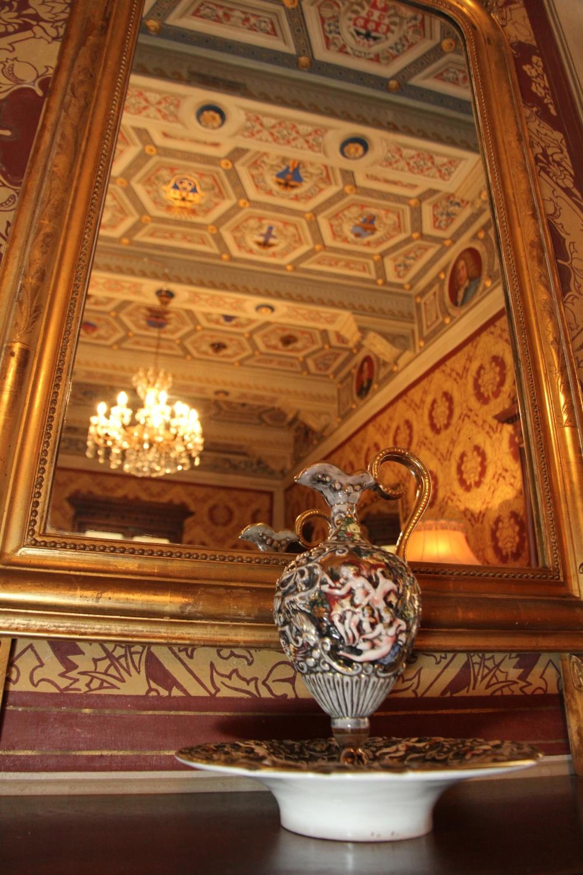 Un miroir avec un cadre doré reflétant une pièce dans laquelle on aperçoit un lustre et un vase décoratif posé sur une table.