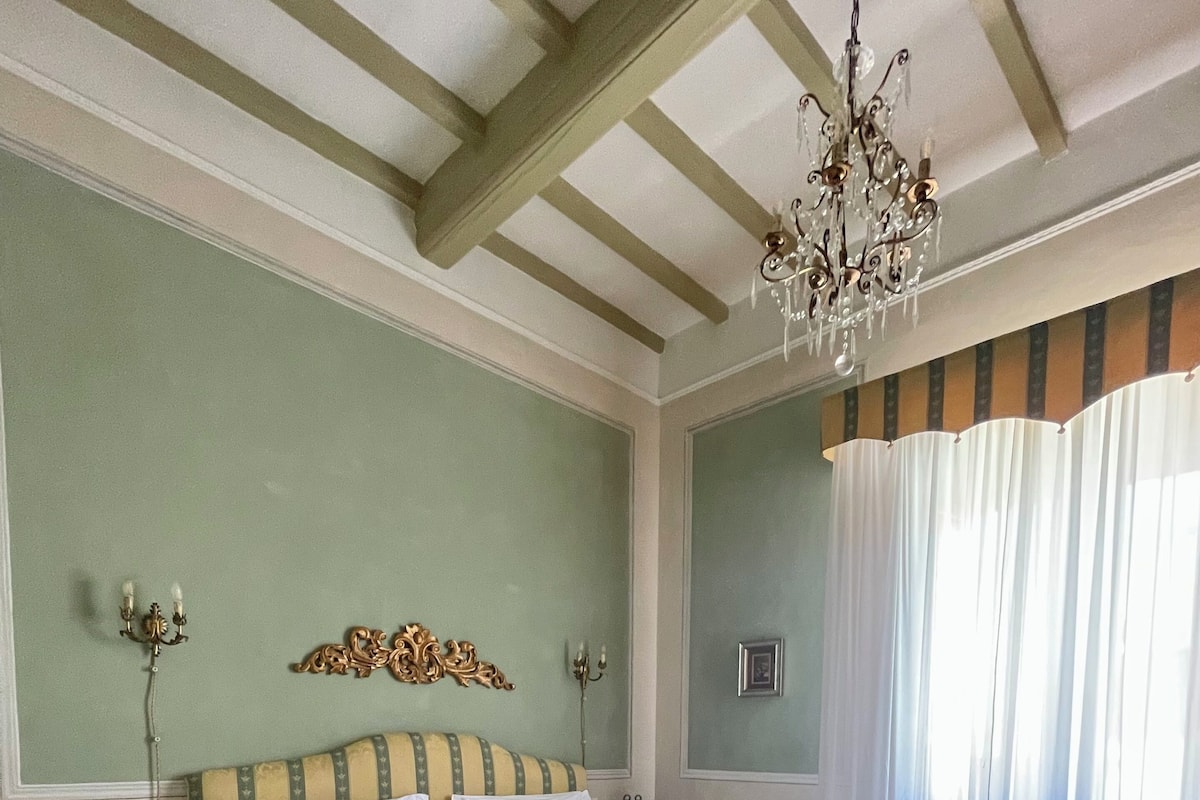 A neatly made bed with a yellow bedspread, a chandelier, and a green wall with a decorative headboard.