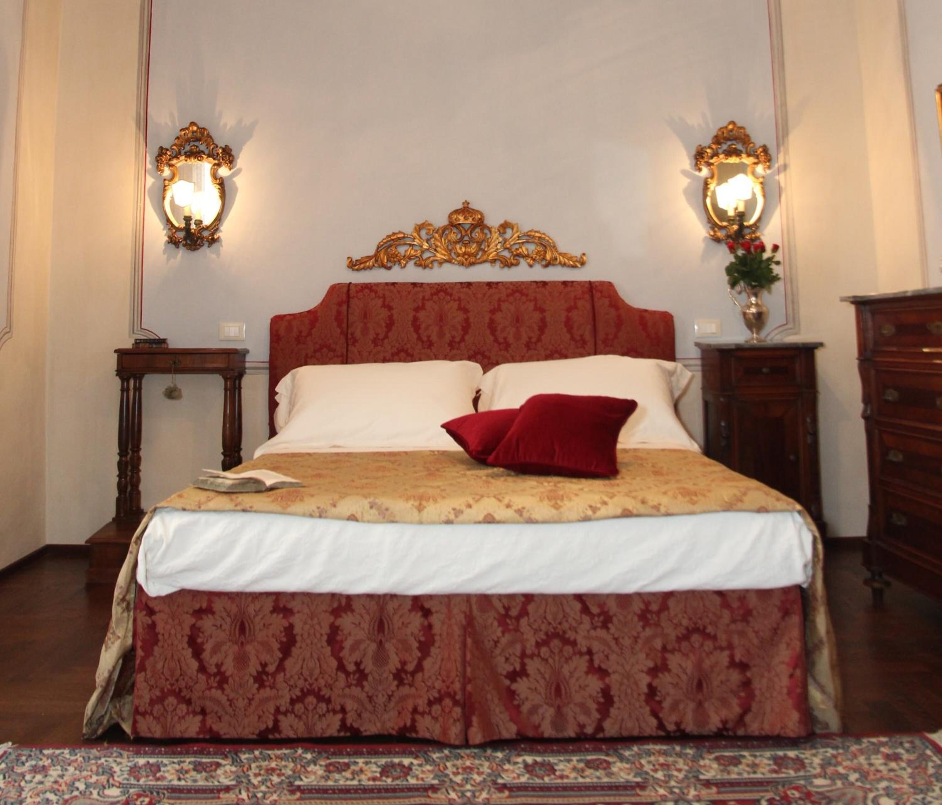A bedroom with a large bed, ornate headboard, two wall sconces, a nightstand, a dresser, and a patterned rug.