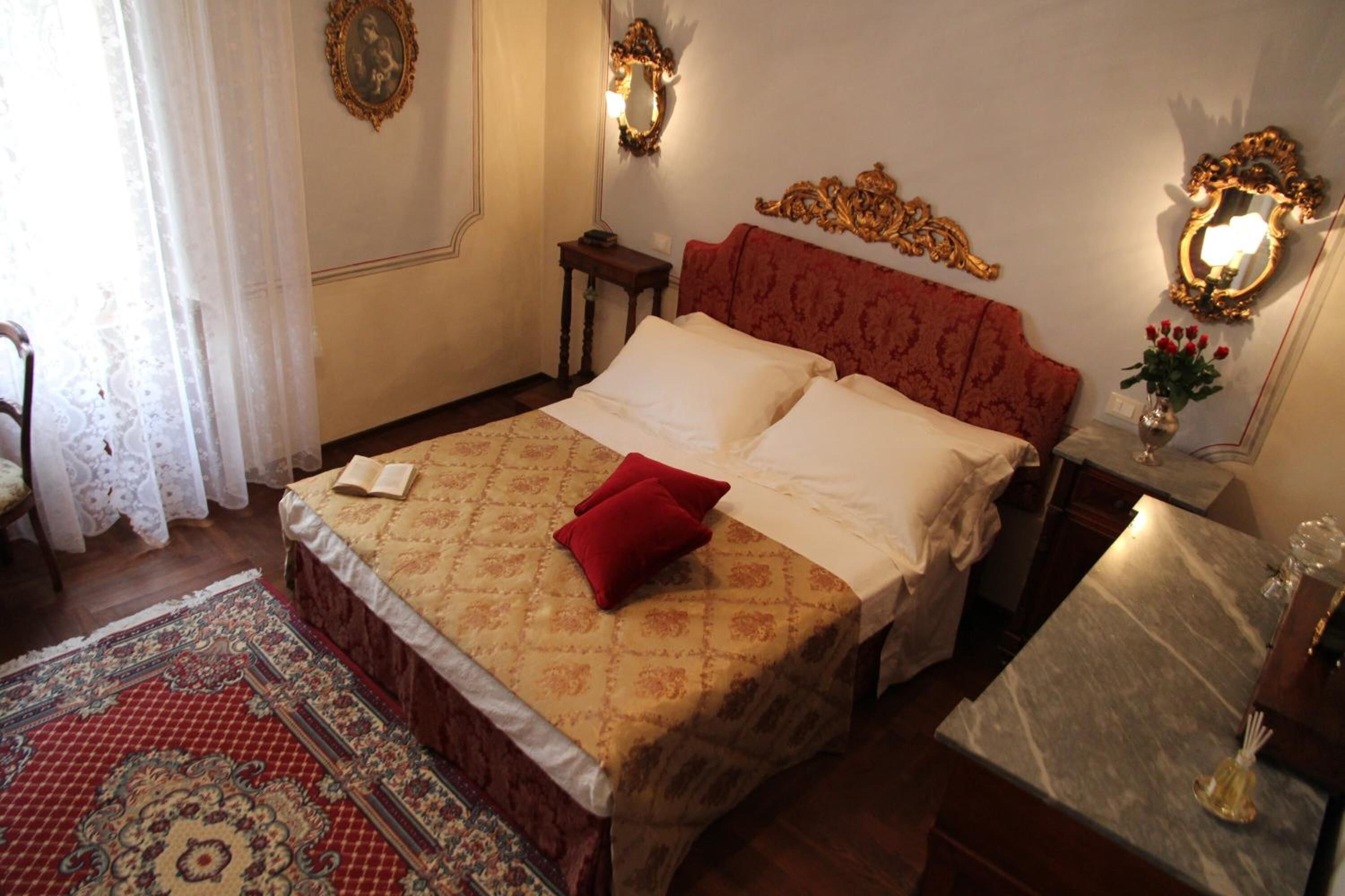 A bedroom with a large bed, a red headboard, two wall sconces, a nightstand, a mirror, and a small table with a vase of flowers.
