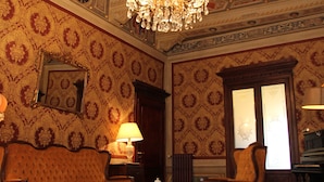 A room with ornate ceiling, a chandelier, a sofa, chairs, a table, and a patterned rug.