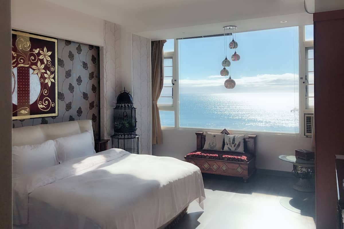 A bedroom with a large bed, a window with a view of the sea, a decorative wall, and a small table.