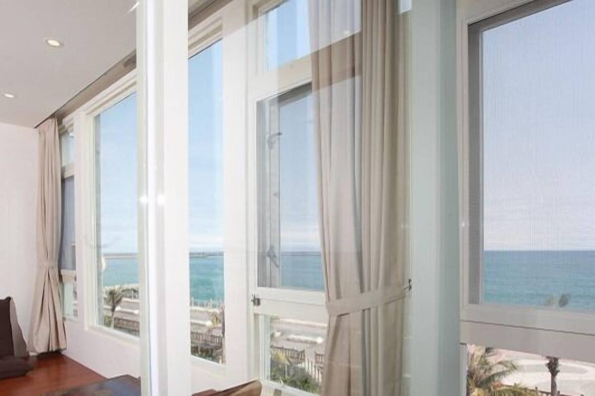 A room with large windows overlooking a beach, a wooden floor, and a dark-colored sofa.