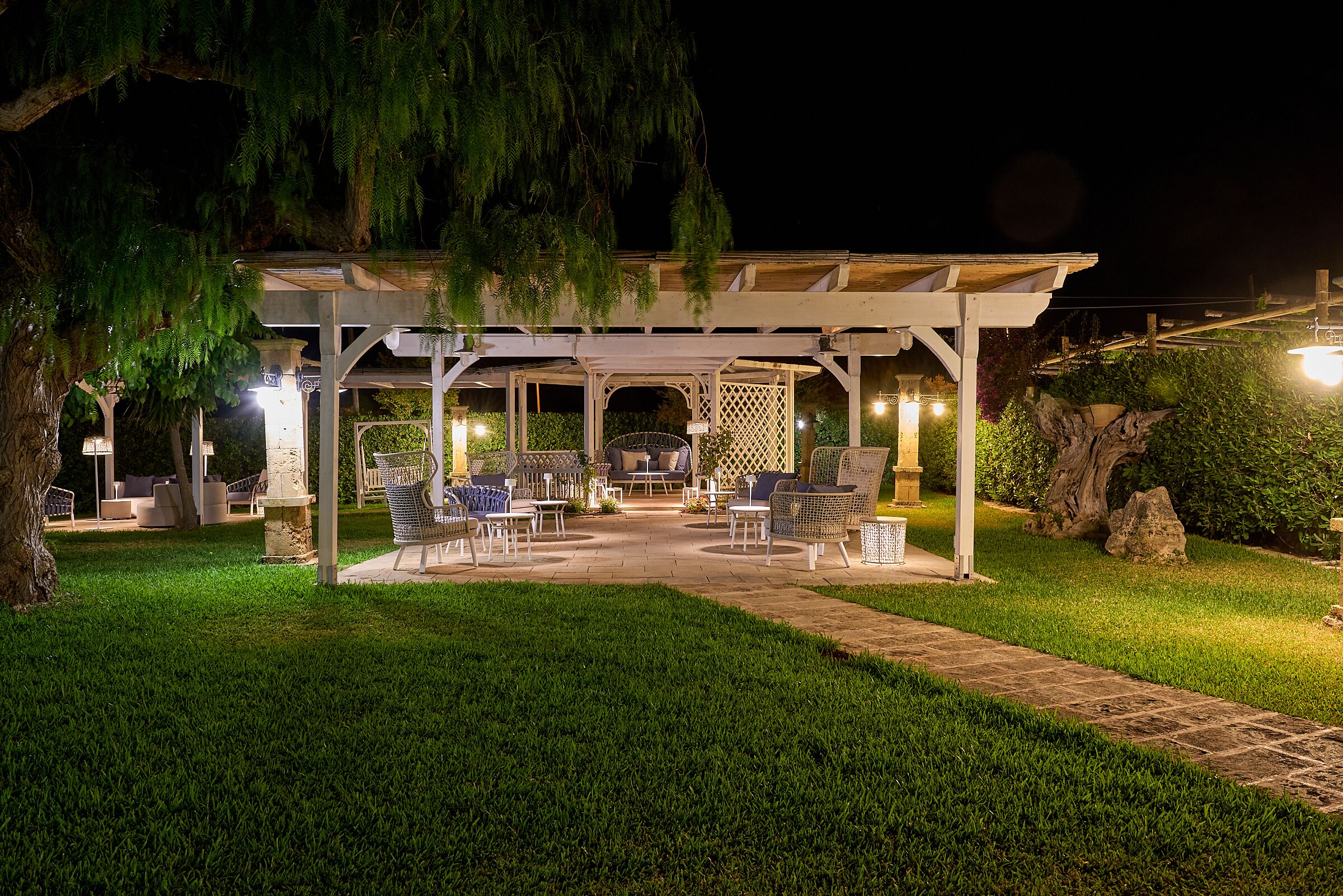 A covered outdoor seating area with chairs and tables, surrounded by well-maintained grass and a stone pathway.