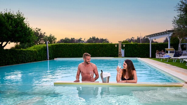 A pool area with a clear view of the water, a couple holding drinks, and a lounge area with chairs and umbrellas. - Montecallini Hotel - Adults Only (Patu)