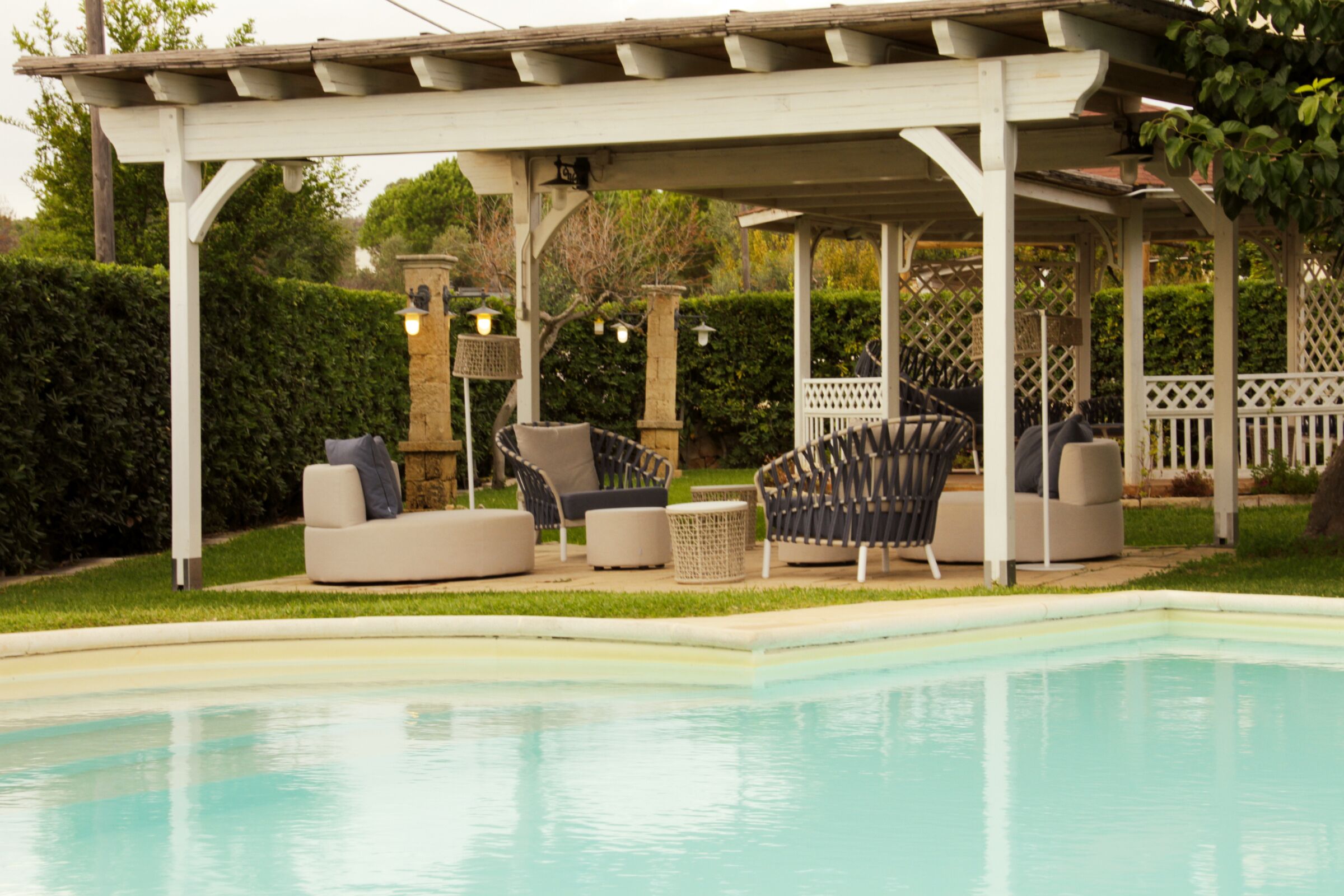 A covered outdoor seating area with a pool, featuring cushioned chairs and a pergola structure.