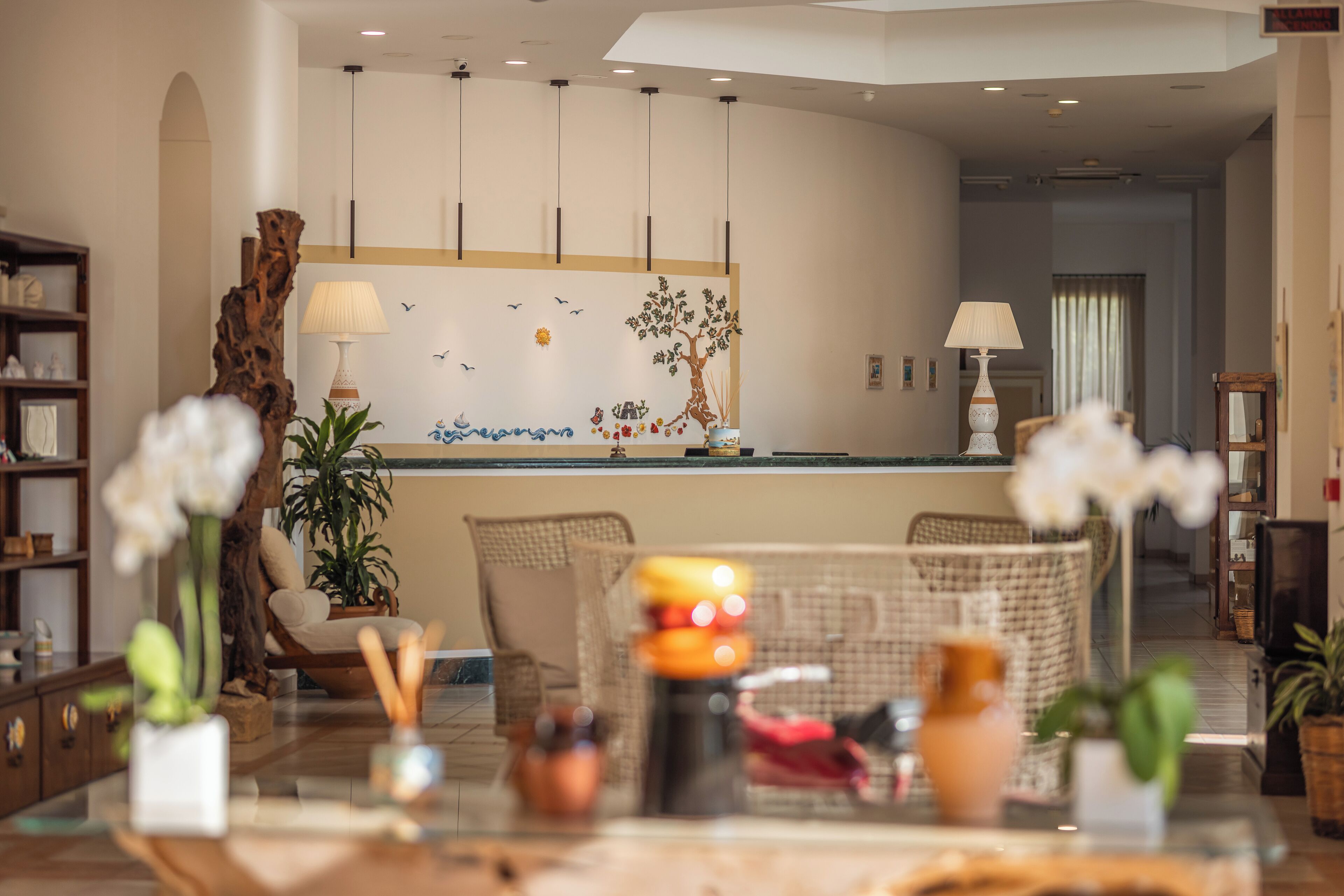 A modern living room with a glass coffee table, wicker chairs, a wooden sideboard, and a large decorative tree mural on the wall.
