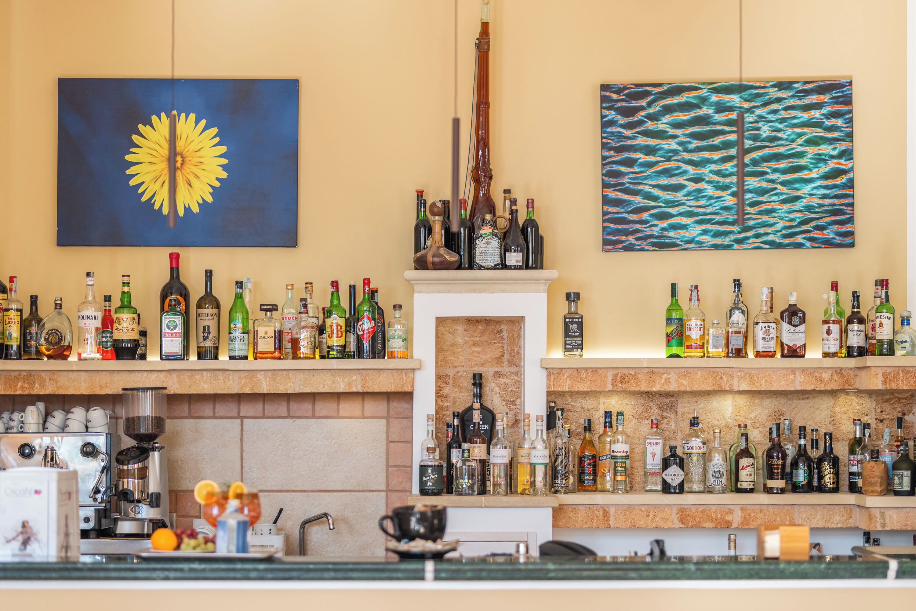 A bar area with shelves of liquor bottles, a coffee machine, and a painting of a sunflower.