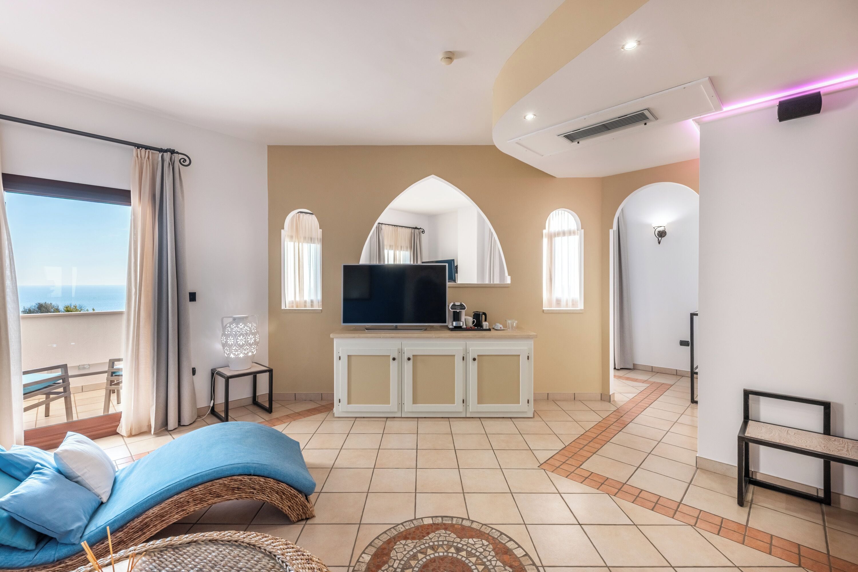 Ein Wohnzimmer mit Fernseher, einer Couch, einem kleinen Tisch und einer Bank. Es gibt einen Balkon mit Meerblick.