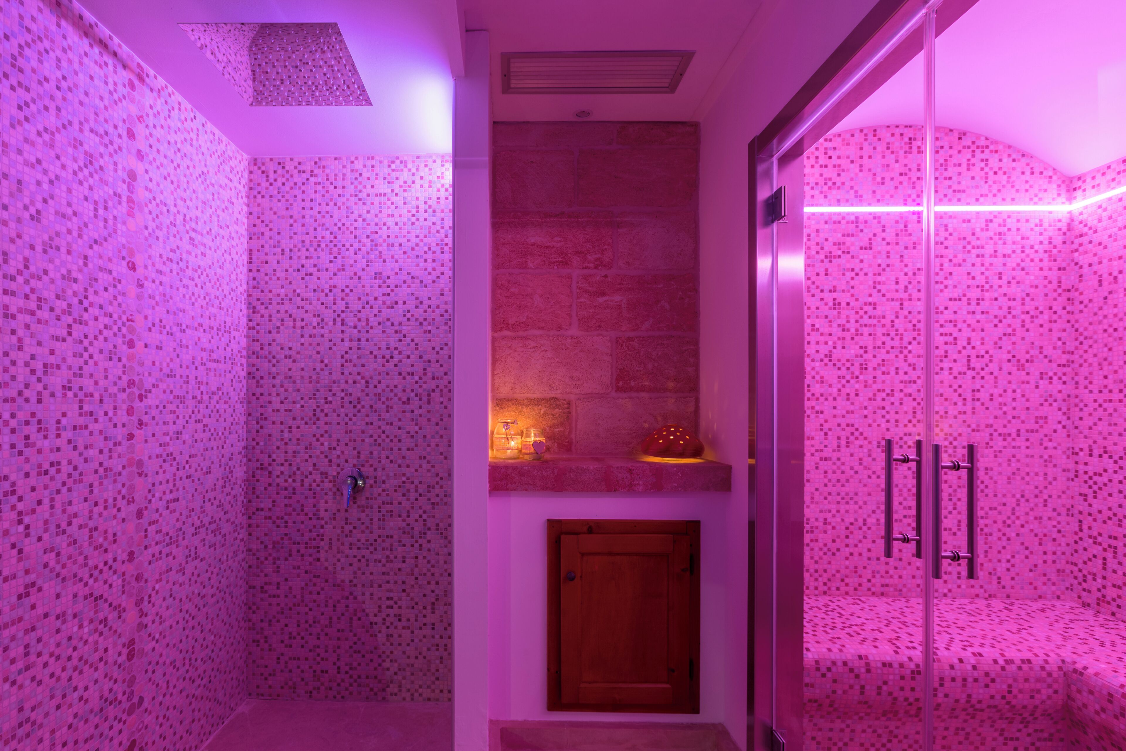 Ett badrum med rosa mosaikplattor, en vägg av natursten, en trädörr och en duschavdelning med glasväggar.