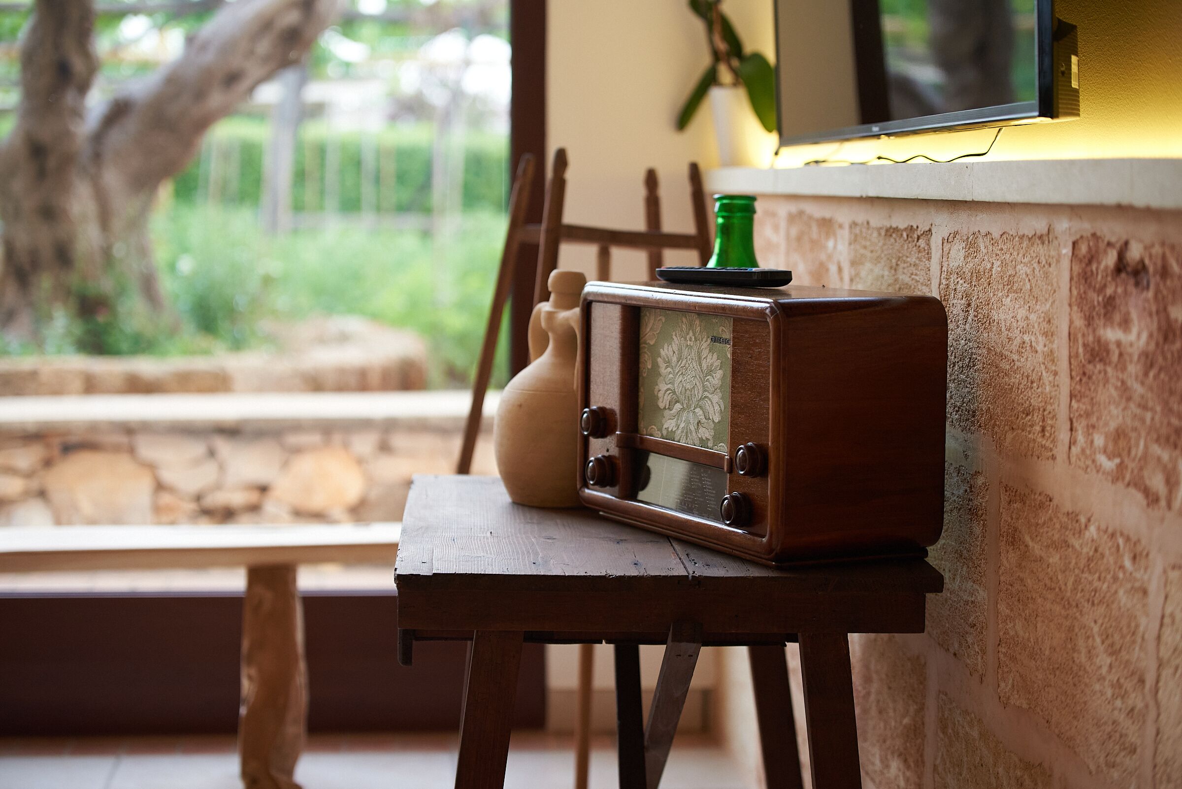 A rustic wooden table with a vintage radio and a green bottle, positioned near a stone wall and a window overlooking greenery.