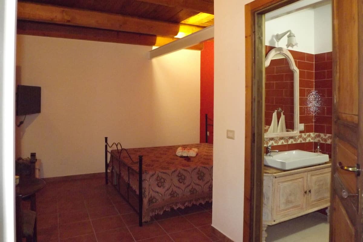 Un dormitorio con cama, techo de madera, suelo de baldosas, mesita de noche, espejo y baño con lavamanos y paredes de azulejos.