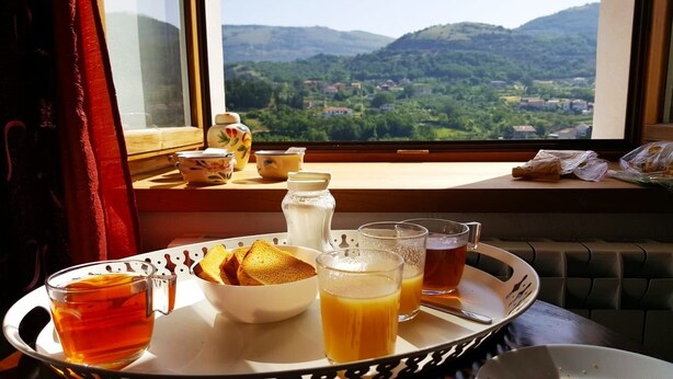 Bandeja de desayuno con té, jugo de naranja y tostadas sobre una mesa junto a una ventana con vistas a colinas verdes.