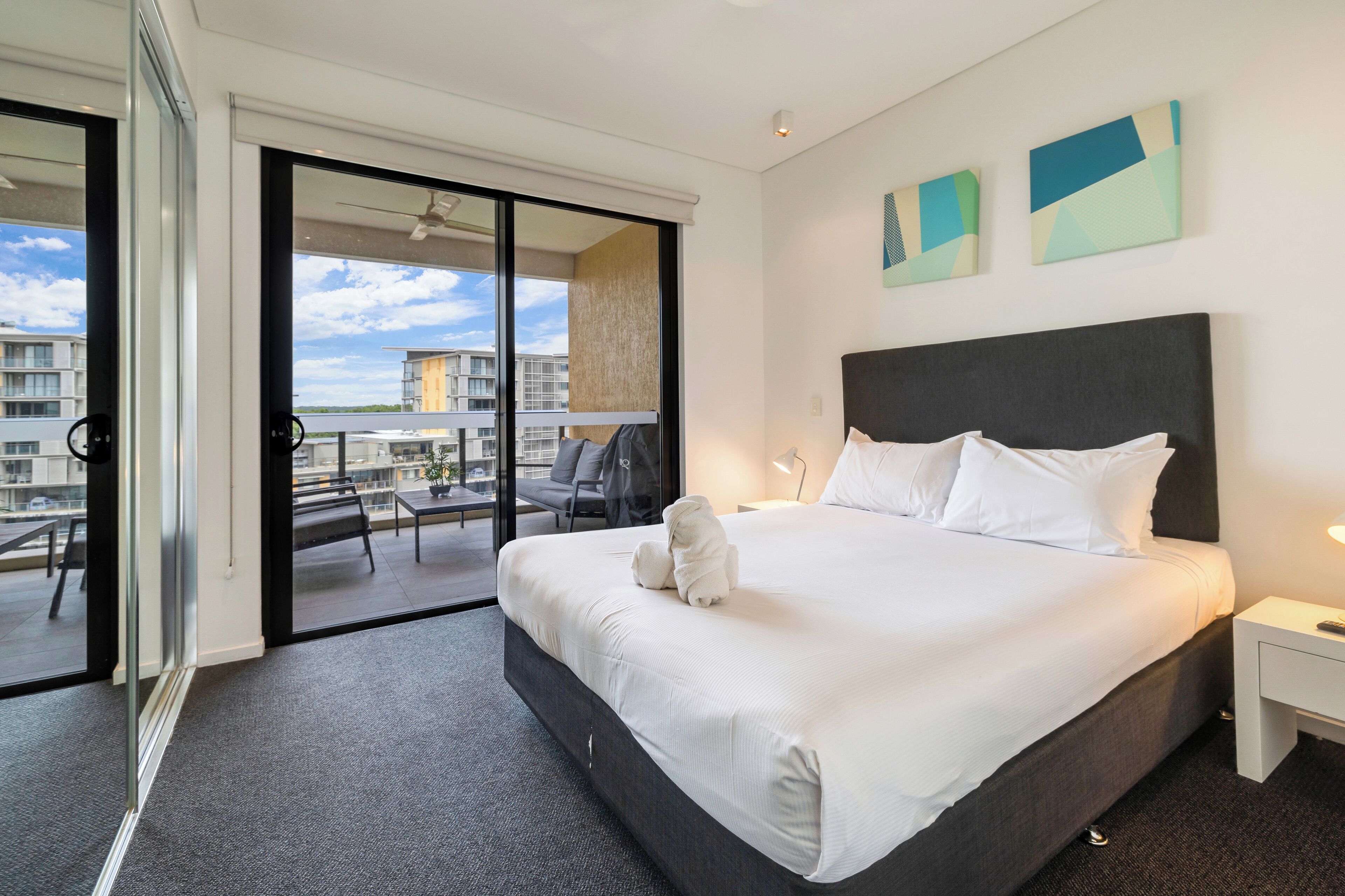 A hotel room with a large bed, a nightstand, a window with a view of the city, and a sliding glass door to an outdoor area.