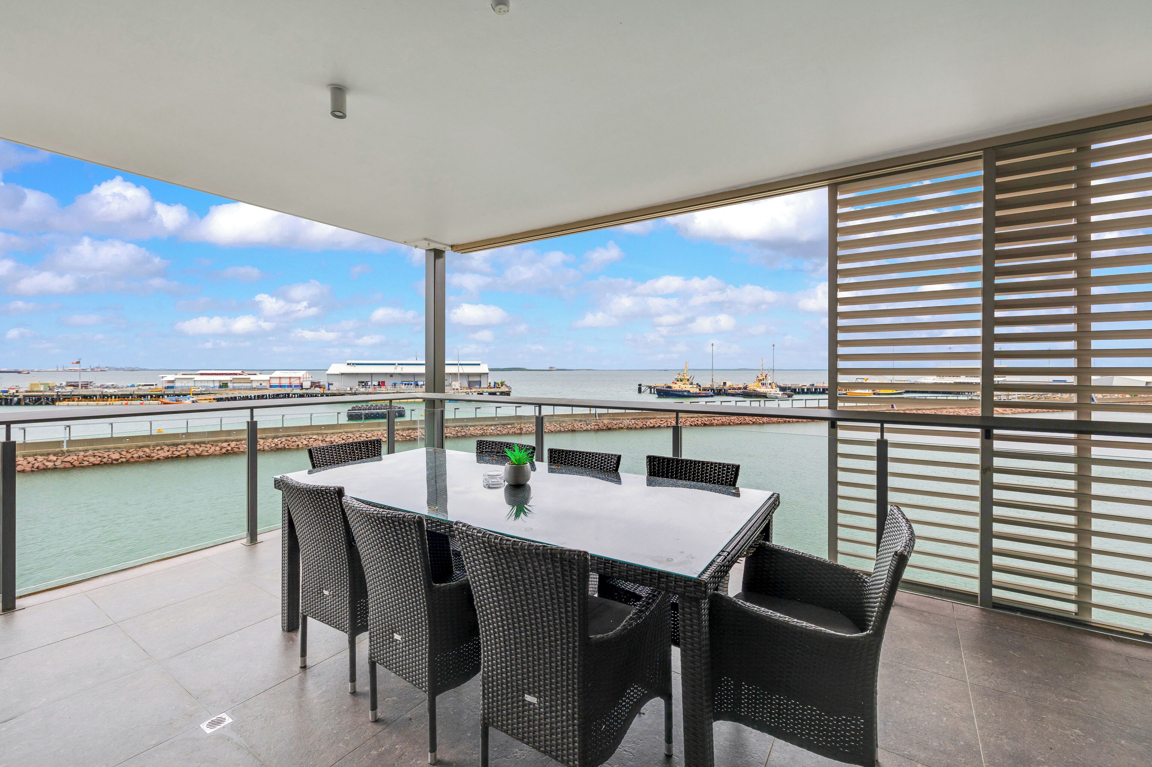 Un balcón con una mesa de vidrio y sillas de mimbre, con vista a un puerto deportivo con barcos y un horizonte urbano distante.