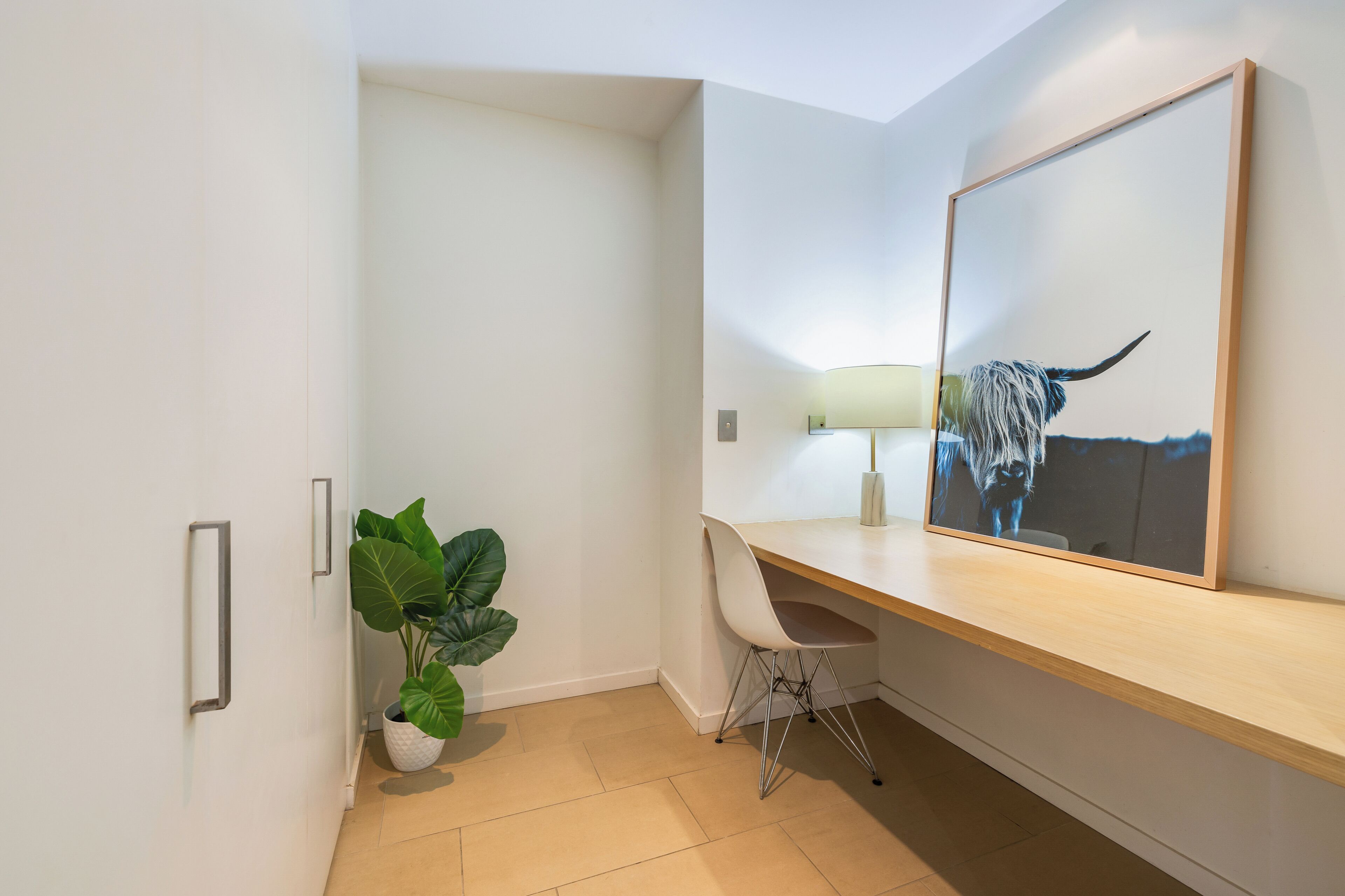 Une chambre d’hôtel moderne avec un bureau en bois, une grande plante en pot, une chaise, une lampe de bureau et une œuvre d’art encadrée représentant un taureau.